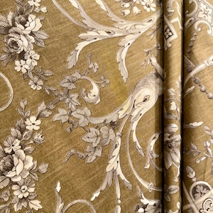 May include: Close-up of a fabric with a repeating floral pattern. The design features white and brown flowers, leaves, and ornate scrollwork against a textured, olive-green background. The fabric is folded, showing the pattern's continuity.