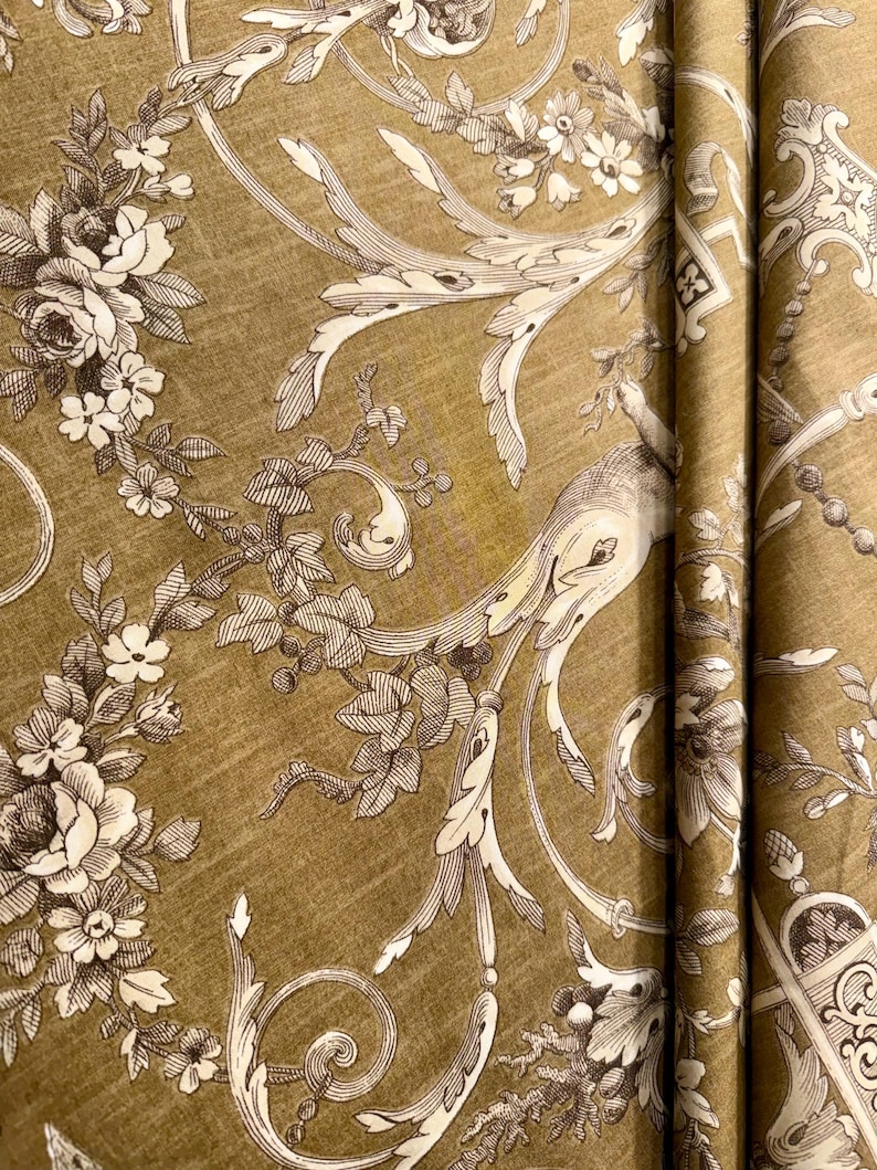 May include: Close-up of a fabric with a repeating floral pattern. The design features white and brown flowers, leaves, and ornate scrollwork against a textured, olive-green background. The fabric is folded, showing the pattern's continuity.