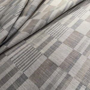Peut inclure: Gros plan sur un tissu plié avec un motif géométrique. Le tissu présente un mélange de carrés, de rectangles et de rayures diagonales dans des tons gris, beige et marron. La texture semble douce et légèrement texturée.
