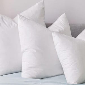 May include: Four white pillows of varying sizes are arranged on a bed. The pillows have a square shape with a pointed top. The pillows are on a light blue sheet, with a gray headboard in the background.