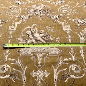 May include: A decorative fabric with a repeating pattern of cherubs, floral elements, and architectural details in shades of beige and brown. A yellow measuring tape is across the fabric, showing measurements in inches.