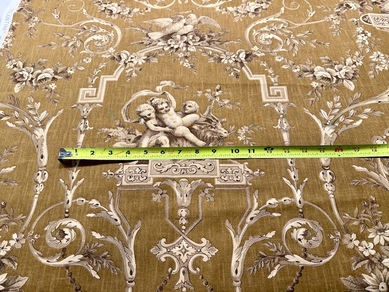 May include: A decorative fabric with a repeating pattern of cherubs, floral elements, and architectural details in shades of beige and brown. A yellow measuring tape is across the fabric, showing measurements in inches.