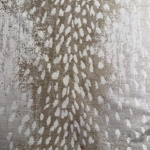 May include: Close-up of a textured fabric with a pattern of white, irregular shapes against a light brown and silver background. The fabric has a distressed, mottled appearance, with a subtle sheen.