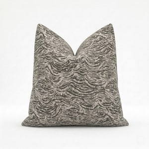 Puede incluir: Cojín decorativo cuadrado con una paleta de colores neutros. El cojín presenta un patrón repetitivo en forma de olas en tonos grises y blanquecinos. El cojín es de forma cuadrada y probablemente se utiliza para la decoración del hogar.