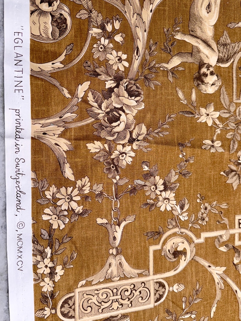 May include: Close-up of a fabric with a detailed floral and cherub design in shades of brown and cream. The fabric features roses, daisies, and scrolling vines. Text on the side reads "EGLANTINE" printed in Switzerland, &copy; MCMXCV.