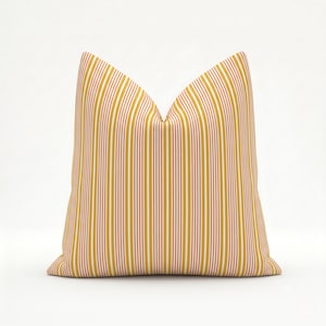 May include: A square throw pillow featuring a vertical striped pattern in shades of mustard yellow, white, and thin rust-colored lines. The pillow has a crisp, structured shape with a pointed top, displaying a repeating linear design across the fabric.