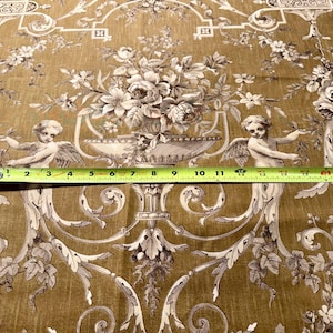 May include: A detailed close-up of fabric with a classic design. The fabric features a repeating pattern of floral arrangements, cherubs, and ornate scrollwork in shades of brown and cream. A measuring tape shows a width of approximately 12 inches.