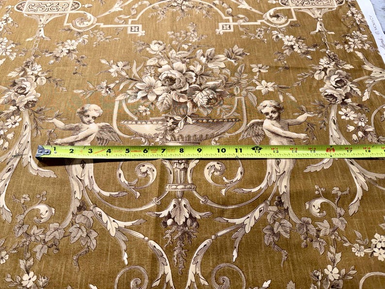 May include: A detailed close-up of fabric with a classic design. The fabric features a repeating pattern of floral arrangements, cherubs, and ornate scrollwork in shades of brown and cream. A measuring tape shows a width of approximately 12 inches.
