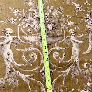 May include: A detailed close-up of a fabric with a classic design. The fabric features a repeating pattern of cherubs, floral arrangements, and ornate scrollwork in shades of brown and cream against a golden-brown background. A measuring tape shows a length of approximately 18 inches.