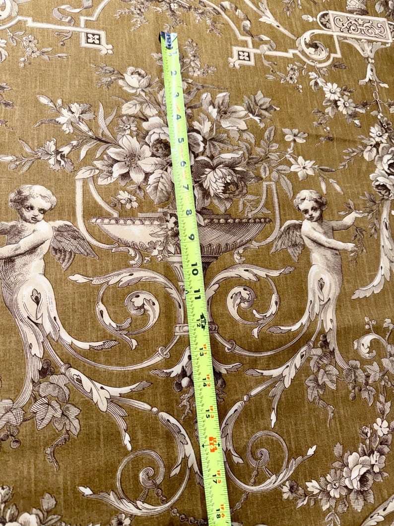 May include: A detailed close-up of a fabric with a classic design. The fabric features a repeating pattern of cherubs, floral arrangements, and ornate scrollwork in shades of brown and cream against a golden-brown background. A measuring tape shows a length of approximately 18 inches.