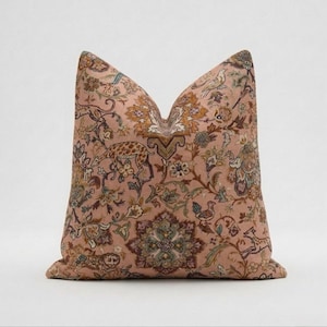 May include: A cushion with a pink backdrop, displaying an intricate design of giraffes, birds, and floral motifs in brown, green, and gold hues. The cushion has a traditional form and a soft, welcoming look.