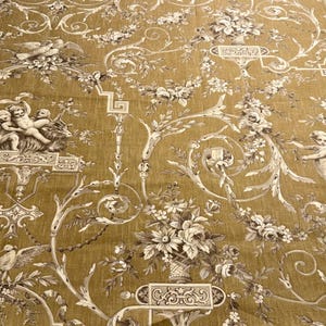 May include: Mustard-colored fabric with a repeating pattern of cherubs, birds, and floral bouquets in shades of white and brown. The fabric has a vintage, classical design, suitable for upholstery or decorative projects.