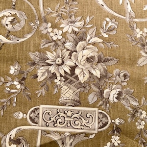 May include: Close-up of a floral pattern on a fabric. The design features a bouquet of flowers in a woven basket, surrounded by scrolling vines and decorative elements in shades of brown and cream against a golden-brown background.