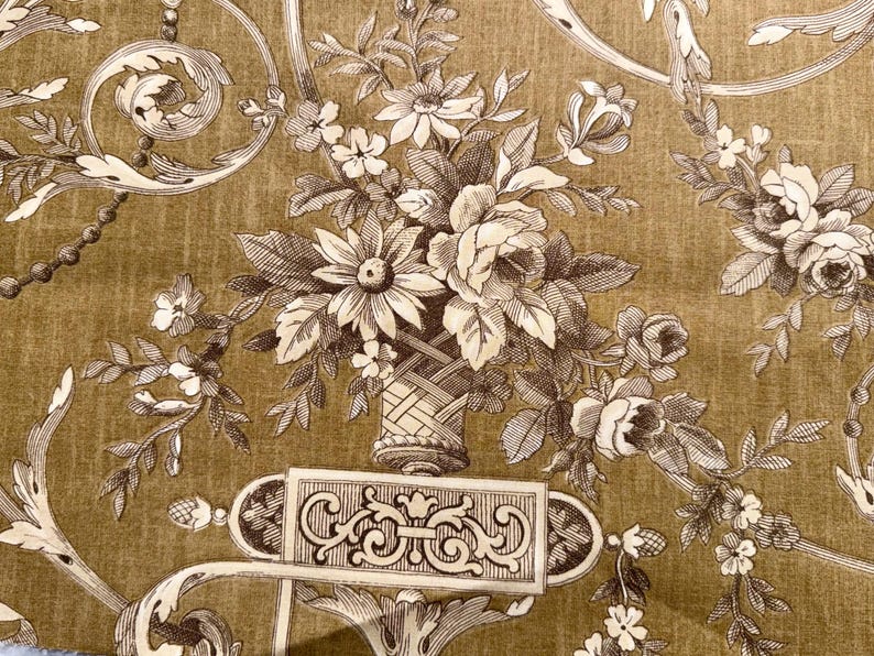 May include: Close-up of a floral pattern on a fabric. The design features a bouquet of flowers in a woven basket, surrounded by scrolling vines and decorative elements in shades of brown and cream against a golden-brown background.