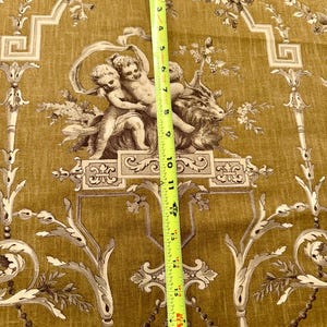 May include: Mustard-colored fabric with a classical design featuring cherubs, floral patterns, and ornate scrollwork. The fabric is measured with a yellow tape measure, showing a length of approximately 18 inches.