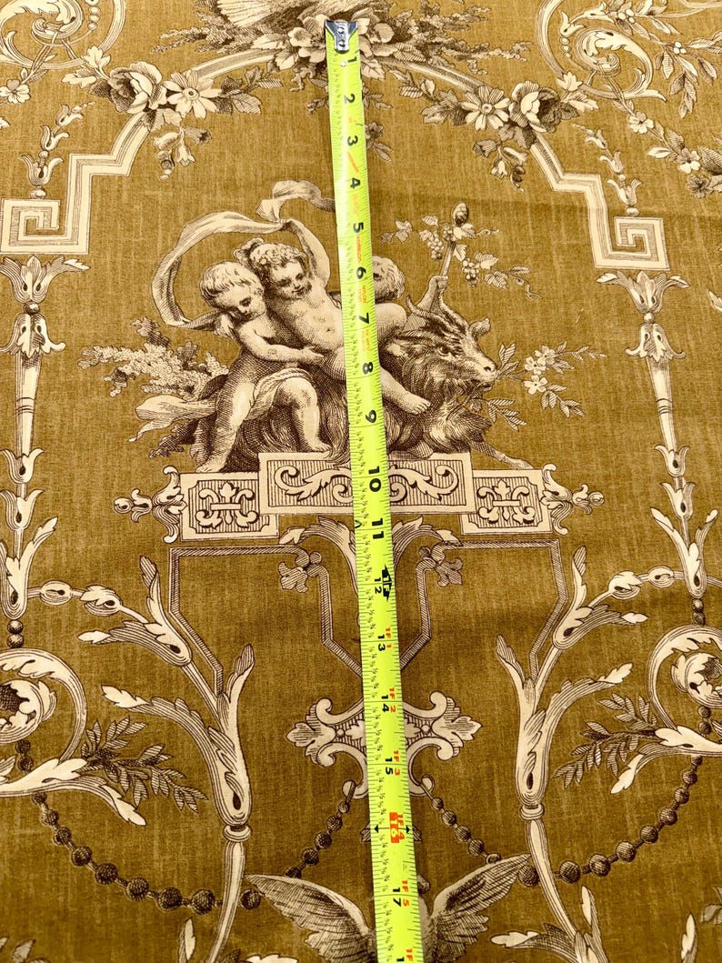 May include: Mustard-colored fabric with a classical design featuring cherubs, floral patterns, and ornate scrollwork. The fabric is measured with a yellow tape measure, showing a length of approximately 18 inches.