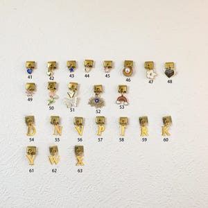 Puede incluir: Una colección de colgantes de metal dorado con varios diseños, incluyendo un corazón, una estrella, una mariposa y un arcoíris. Algunos colgantes presentan letras. Los colgantes se muestran sobre un fondo blanco.