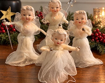 May include: Four vintage-style angel figurines with pale skin, painted facial features, and blonde hair. Each angel wears a white sheer gown with silver trim and translucent wings. Two angels hold gold-colored star wands on thin metal rods.
