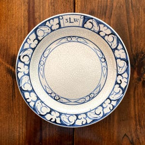 May include: A white ceramic plate with a blue floral design around the rim and a blue inner circle. The plate has the letters "sLw" at the top. The plate has a cracked, aged appearance.