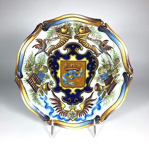 May include: A decorative ceramic plate with a scalloped edge featuring a central coat of arms with a crown and blue ribbon on a tan background. The design includes cornucopias, pan flutes, grapes, and winged motifs in blue, gold, and green.