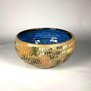 May include: A handmade ceramic bowl with a textured, tan exterior adorned with colorful, abstract patterns. The interior is a vibrant blue. The bowl is round and has a wide opening, suitable for serving or display.