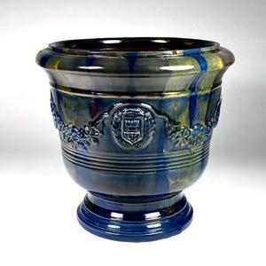 May include: A glazed ceramic planter featuring a deep blue and olive green drip finish. The exterior is decorated with a raised relief pattern of floral garlands and a central crest emblem. The vessel has a wide rim and a pedestal base.