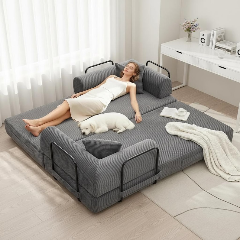 May include: A gray convertible sofa with black metal armrests. A person in a white dress and a white dog are resting on the sofa. A white blanket and a book with a cup are on the sofa.