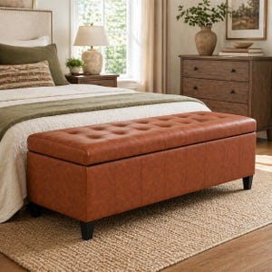 May include: A rectangular, brown leather storage ottoman with a tufted top. The ottoman has black legs and is placed at the foot of a bed. The bed has a white comforter, green throw, and decorative pillows. The room has a neutral color palette.