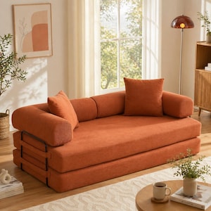 Puede incluir: Un sofá cama naranja con dos cojines y cojines de apoyo a juego. El sofá tiene una estructura de metal negro. La habitación está decorada con una lámpara de pie, obras de arte y una pequeña mesa auxiliar. El sofá mide unos 200 cm de largo.