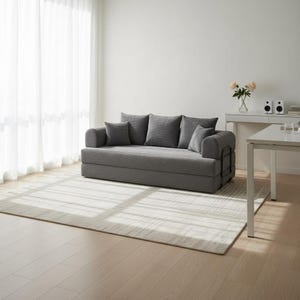 May include: A gray convertible sofa with several matching pillows. The sofa sits on a light-colored rug in a room with a white wall and sheer curtains. The sofa has a simple, modern design.