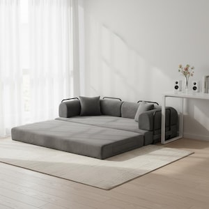 May include: A gray convertible sofa bed with a modern design. The sofa is partially unfolded, revealing a flat sleeping surface. It has gray cushions and pillows, and black metal armrests. The sofa sits on a light-colored rug in a well-lit room.