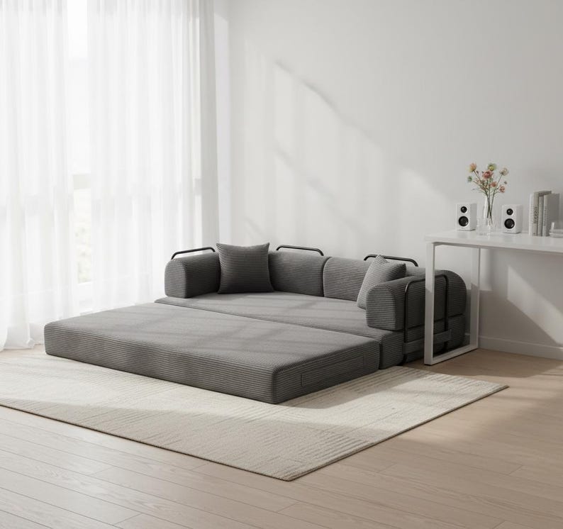 May include: A gray convertible sofa bed with a modern design. The sofa is partially unfolded, revealing a flat sleeping surface. It has gray cushions and pillows, and black metal armrests. The sofa sits on a light-colored rug in a well-lit room.