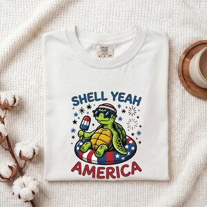 Op de afbeelding: Een wit t-shirt met een afbeelding van een groene schildpad met een zonnebril en een hoofdband, zittend in een rood, wit en blauwe zwemband terwijl deze een ijsje vasthoudt. De tekst luidt SHELL YEAH AMERICA, omringd door sterren en vuurwerkiconen.