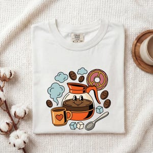 Può includere: Una t-shirt bianca con una stampa grafica raffigurante una caffettiera arancione sorridente, una piccola tazza arancione con un cuore rosso, una ciambella rosa, chicchi di caffè con faccine, nuvole di vapore azzurre, zollette di zucchero e un cucchiaio.