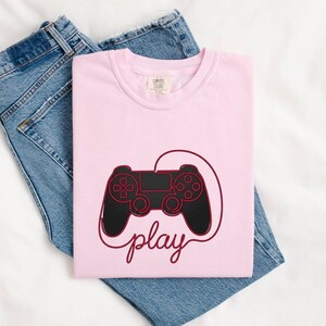 Può includere: Una t-shirt rosa chiaro con una grafica nera e rosso scuro di un controller per videogiochi con un cavo che forma la parola play in corsivo. La maglietta ha un girocollo ed è appoggiata sopra un paio di jeans in denim blu.