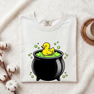 Puede incluir: Una camiseta blanca con un gráfico de un patito de goma amarillo flotando en un caldero negro lleno de líquido verde y burbujas. La camiseta tiene cuello redondo y una etiqueta de la marca Comfort Colors impresa en el interior.