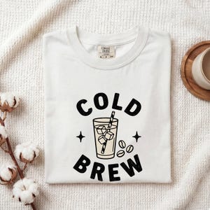 Può includere: Una t-shirt bianca a maniche corte con una stampa grafica nera che recita COLD BREW. Il design include un'illustrazione centrale di un bicchiere riempito con cubetti di ghiaccio e una cannuccia, circondato da piccole stelle e chicchi di caffè.