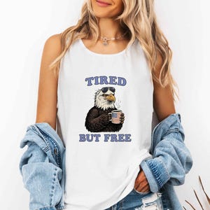 May include: A white sleeveless tank top featuring a graphic of a bald eagle wearing sunglasses and holding a steaming mug with an American flag pattern. The text TIRED BUT FREE is printed in blue letters above and below the eagle illustration.