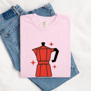 May include: A light pink t-shirt featuring a graphic of a red moka pot coffee maker with a black handle and lid, surrounded by four small red four-pointed star icons. The shirt is folded and placed on top of a pair of blue denim jeans.