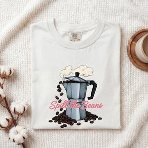 Puede incluir: Una camiseta blanca con una ilustración gráfica de una cafetera italiana plateada con vapor saliendo de la parte superior y granos de café derramándose desde la base. El texto "Spill the Beans" está impreso en cursiva rosa en el frente.