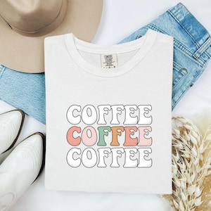 Może przedstawiać: Biały t-shirt z krótkim rękawem z napisem COFFEE wydrukowanym trzy razy w stosie, falowaną czcionką w stylu retro. Górny i dolny wiersz są białe z czarnymi konturami, natomiast środkowy wiersz zawiera litery w kolorach pastelowego różu, zieleni i pomarańczu.