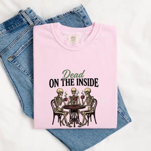 May include: A light pink t-shirt featuring a graphic of three skeletons sitting at a round table drinking from red mugs. The text above the skeletons reads Dead ON THE INSIDE in green and black lettering. The shirt is folded over blue denim jeans.