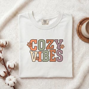 May include: A white short-sleeved t-shirt featuring the text COZY VIBES in a multi-colored block font with small paw prints interspersed. The shirt has a Comfort Colors brand label at the inner neckline and is made of a solid white fabric.