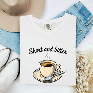 Puede incluir: Una camiseta blanca de manga corta con un gráfico de una taza de café sobre un platillo con una cuchara y el texto negro "Short and bitter" sobre la taza. La camiseta tiene una etiqueta de la marca Comfort Colors en el interior del cuello.