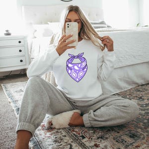May include: A white long-sleeve sweatshirt featuring a central graphic of a purple bandana with a white paisley pattern. The sweatshirt has a crew neckline and ribbed cuffs, providing a clean look that highlights the vibrant purple bandana design.