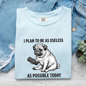 May include: A light blue t-shirt featuring a black and white illustration of a pug sitting and holding a television remote. The text above the pug reads I PLAN TO BE AS USELESS and the text below the pug reads AS POSSIBLE TODAY. The shirt has a Comfort Colors label.