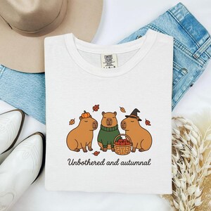 Könnte beinhalten: Ein weißes T-Shirt mit einer Grafik von drei Capybaras, eines mit einem Kürbis, eines in einem grünen Pullover und eines mit einem Hexenhut, das einen Korb mit Äpfeln hält, umgeben von Herbstblättern und dem Text Unbothered and autumnal.