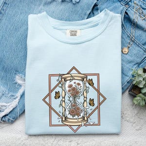 May include: A light blue t-shirt with a graphic print featuring a tan hourglass filled with orange flowers and surrounded by three butterflies. The design is framed by a brown diamond shape. The shirt has a Comfort Colors brand label at the neck.