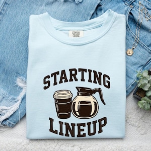 May include: A light blue t-shirt with a dark brown graphic print featuring a coffee cup and a coffee pot. The text above and below the graphic reads "STARTING LINEUP" in a bold, dark brown font. The shirt has a Comfort Colors brand label.