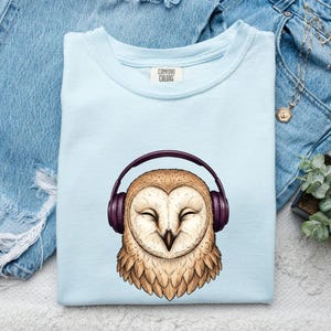 May include: A light blue t-shirt featuring a graphic illustration of a tan barn owl with its eyes closed, wearing dark purple over-ear headphones. The inner neck label displays the text Comfort Colors in black lettering on a white background.